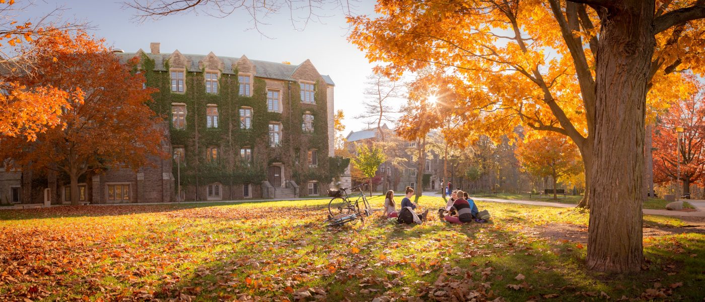 A group of students sit on the grass at McMaster in the fall. Orange, yellow and red trees and leaves are all around them.