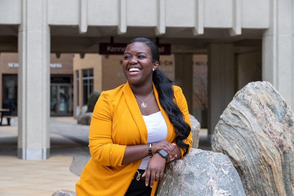 Nicole Agyei-Odame poses for a portrait in front of Togo Salmon Hall. She leans on a rock installation and looks up and away from the camera. 