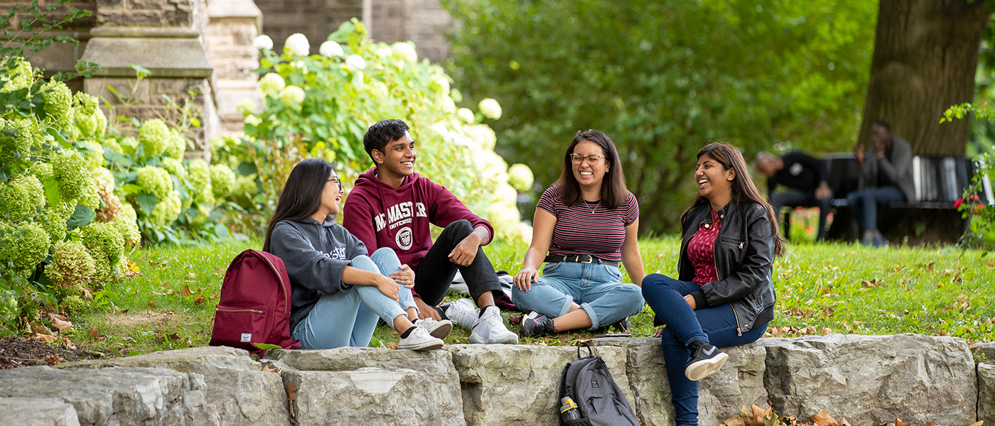 Four McMaster students sit on stone pillars on campus. The grass and bushes behind them are green and it is a warm, sunny summer day.